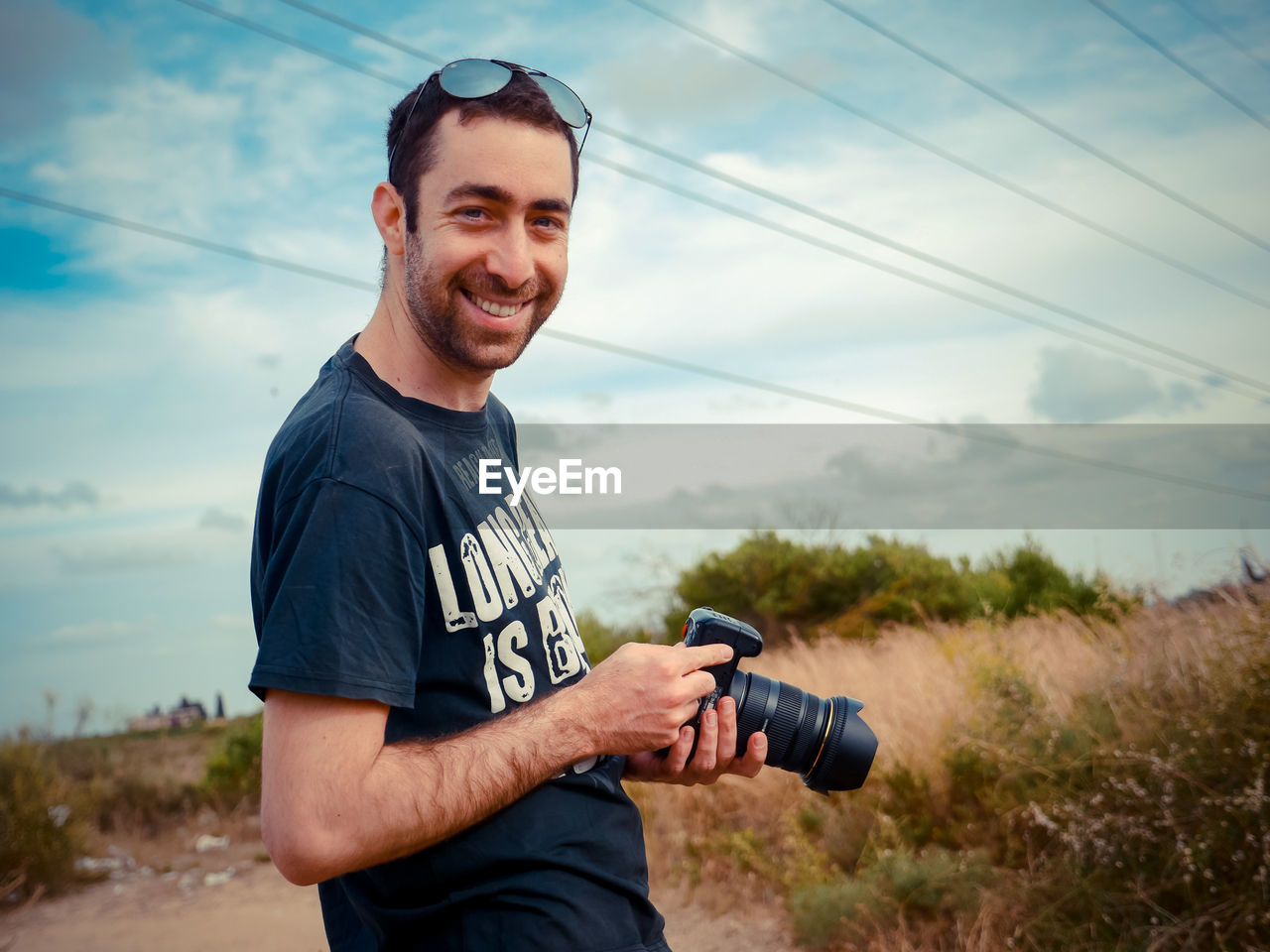 Smiling man holding camera while standing against sky