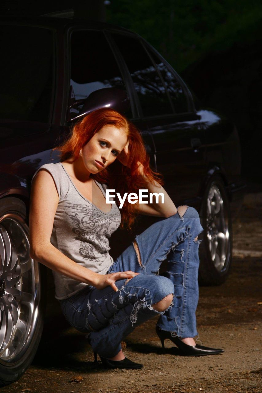 Portrait of young woman crouching by car on dirt road at night