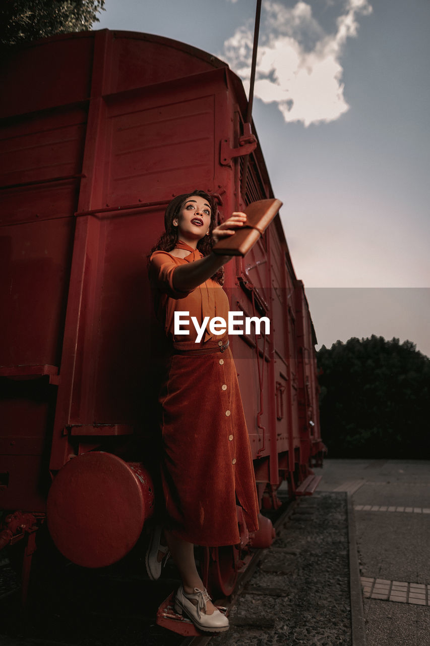 Young female with dark curly hair in beret wearing terracotta clothes in vintage style in back lit standing on step of car train painted in terracotta color and holding handrail while looking away flirty