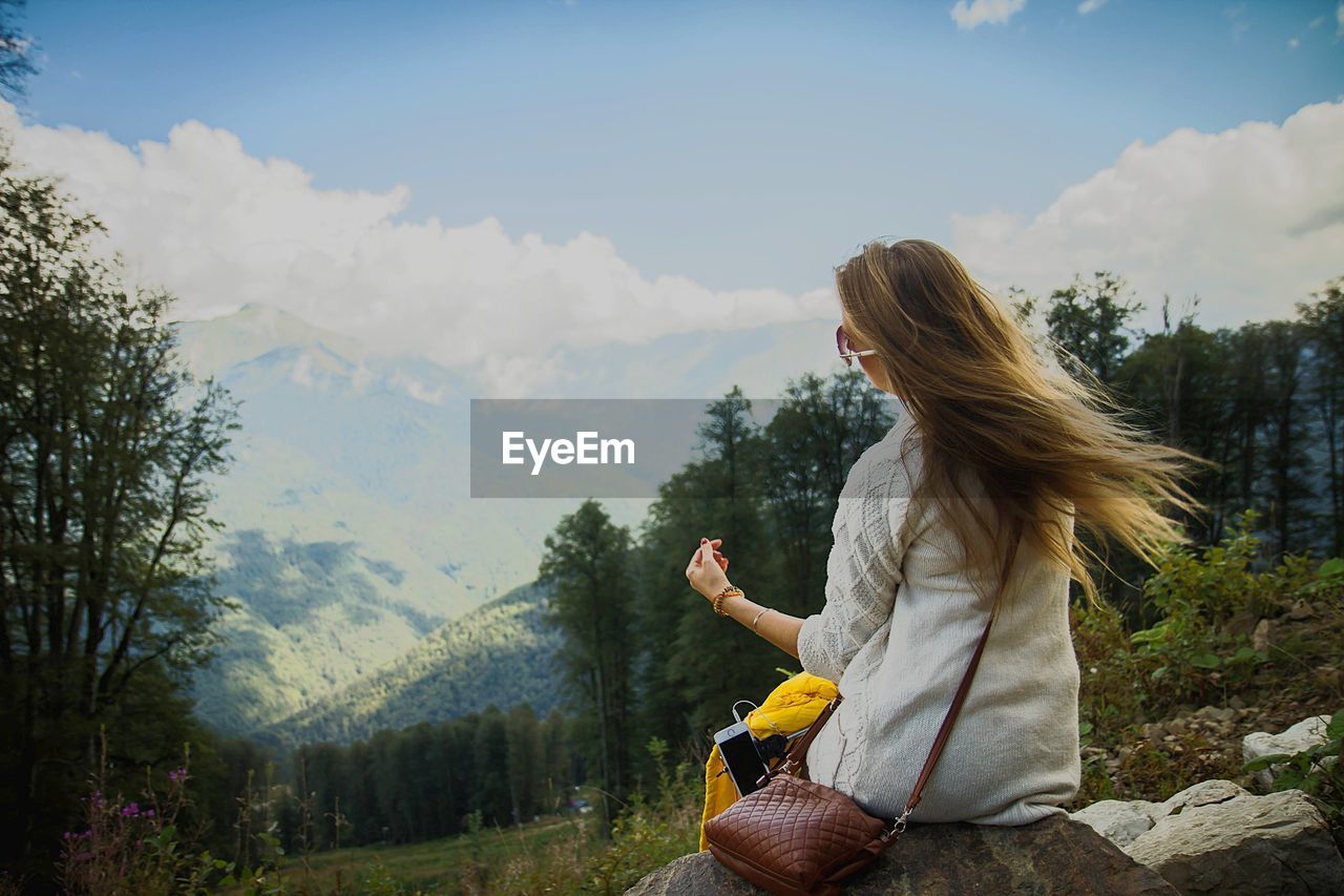 Rear view of woman sitting on rock against sky in forest