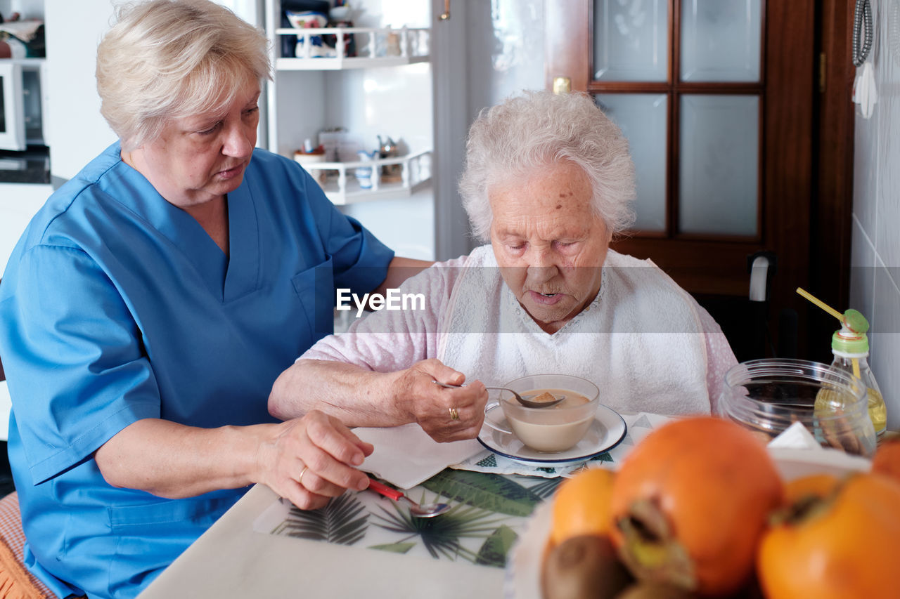 Nurse in uniform sitting near aged woman in wheelchair eating tasty soup at table with food in light kitchen at home