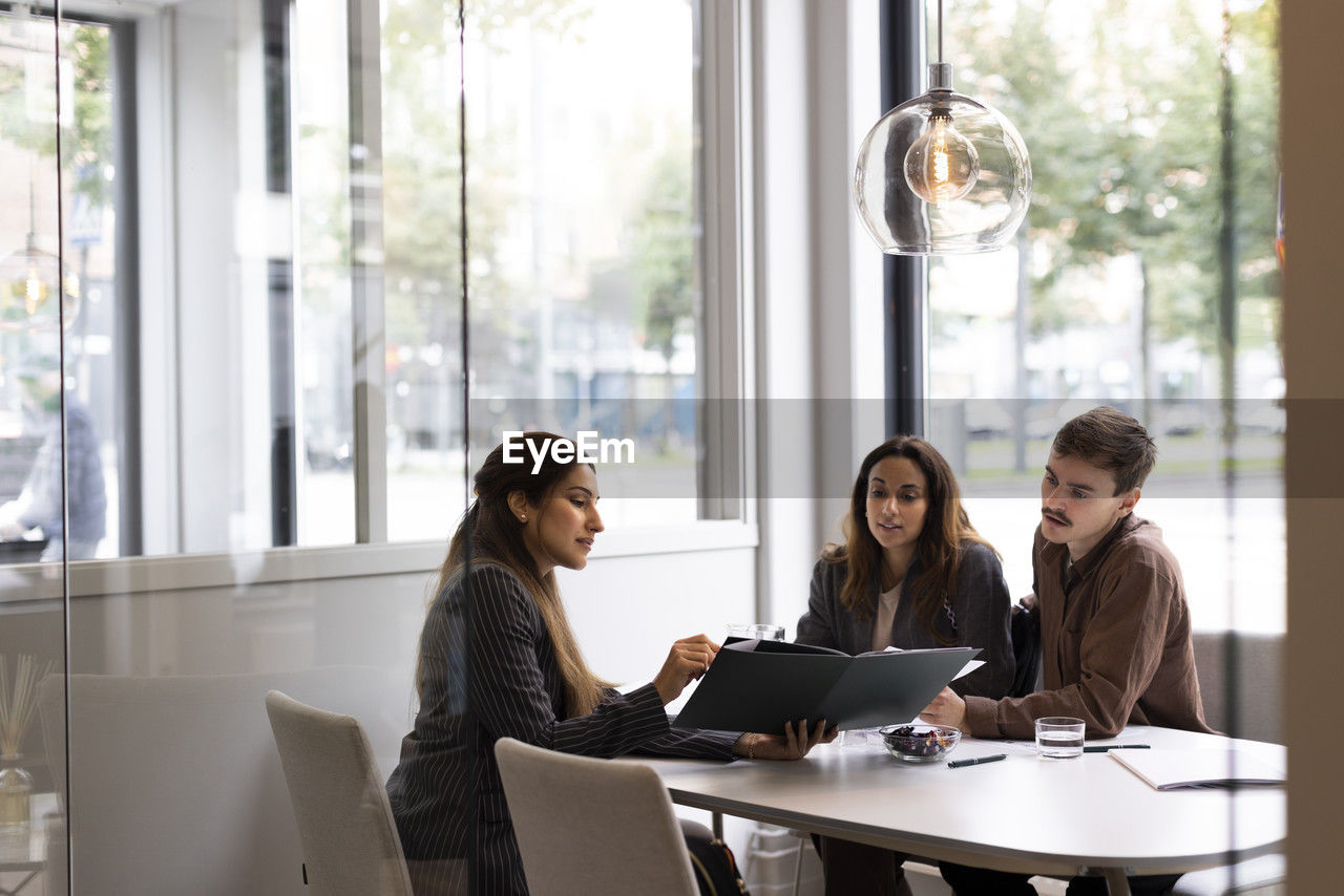 Female real estate agent showing floor plan to young customers while sitting at desk in office