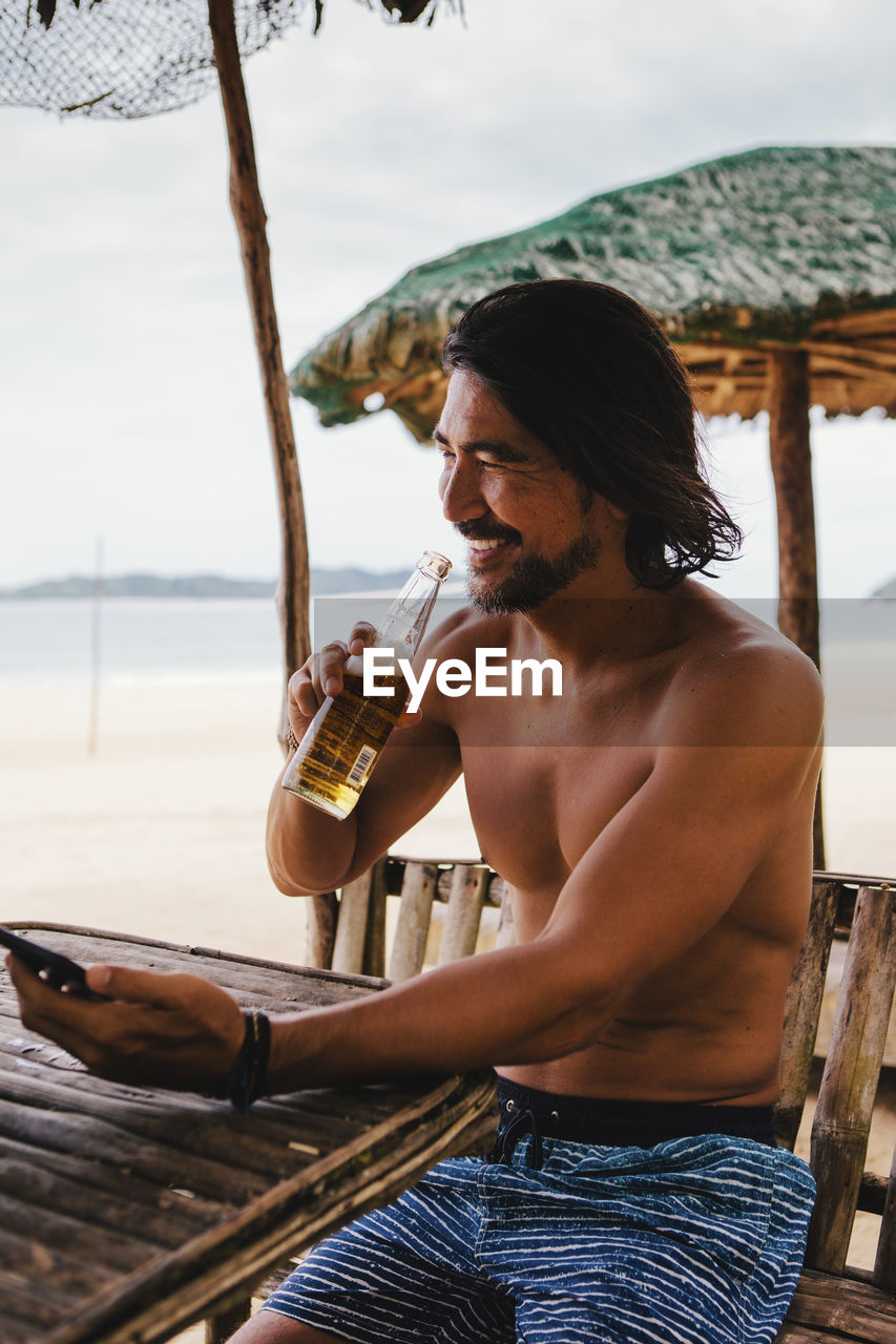 Shirtless man having beer while using mobile phone at beach