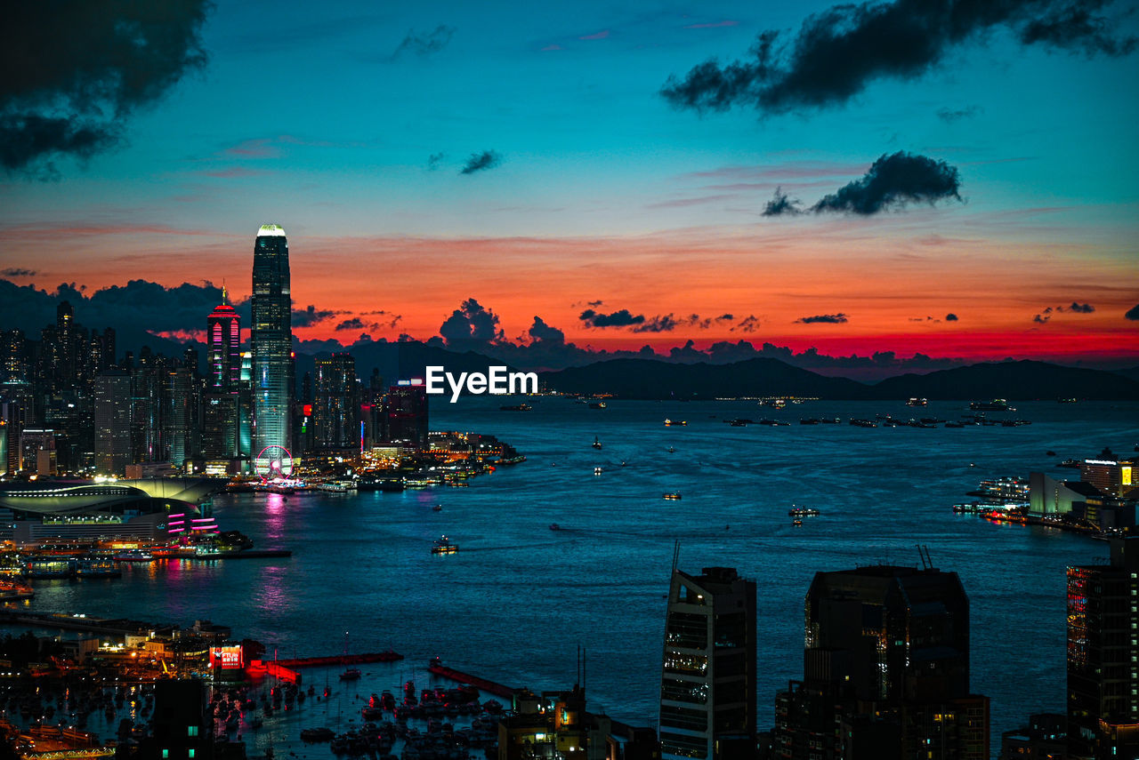 Illuminated city by sea against sky during sunset