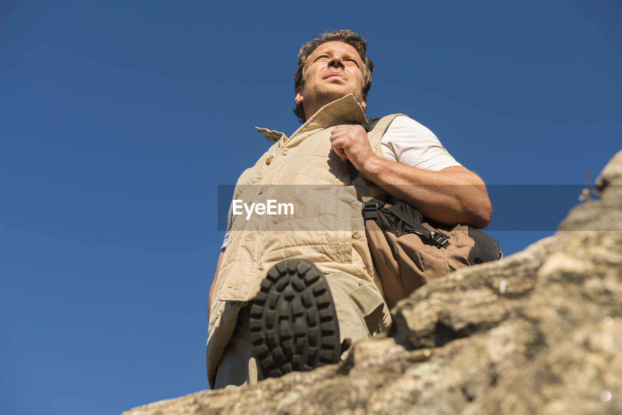 Low angle view of male hiker standing on rock against clear blue sky