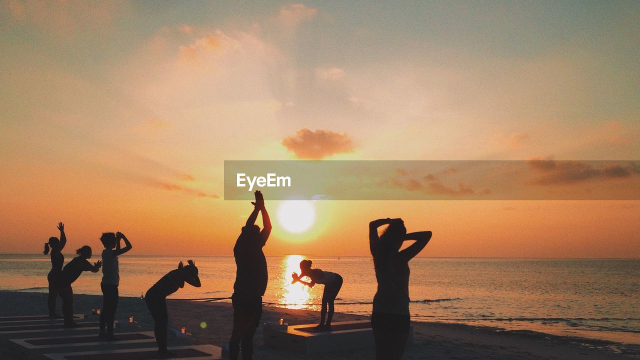 Silhouette women exercising on beach by sea against sky during sunset