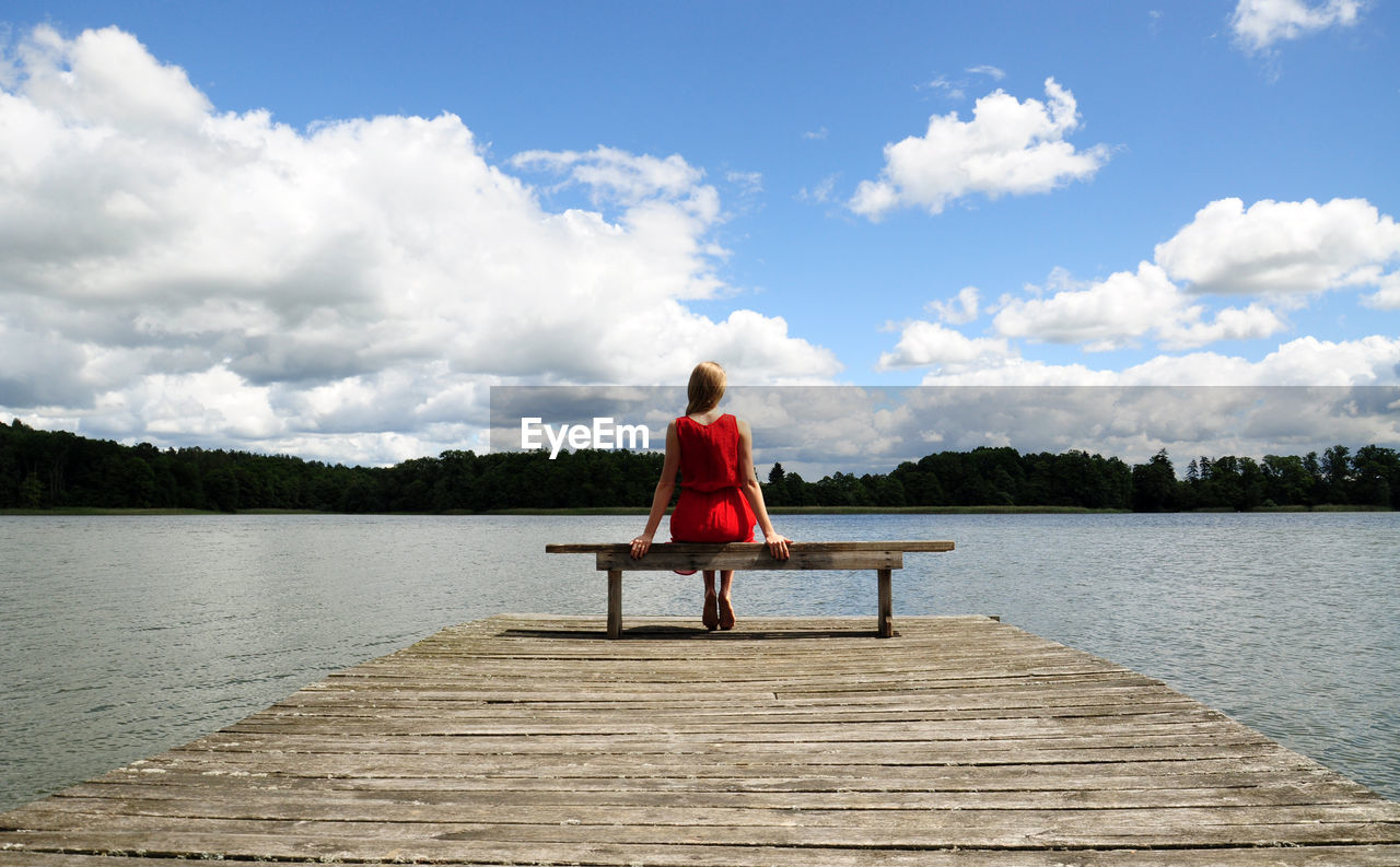 Rear view of woman sitting on pier over lake against cloudy sky