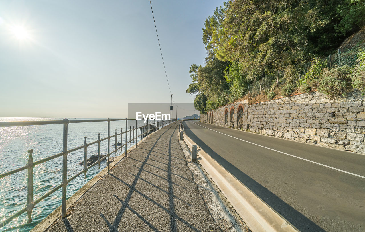 sky, water, nature, sea, walkway, sunlight, transportation, railing, coast, the way forward, day, beauty in nature, tree, plant, land, scenics - nature, beach, diminishing perspective, tranquility, architecture, road, outdoors, no people, tranquil scene, travel, vanishing point, sunny, boardwalk, horizon, horizon over water, travel destinations, shore, clear sky, built structure, footpath, city, idyllic, vacation, street, tourism