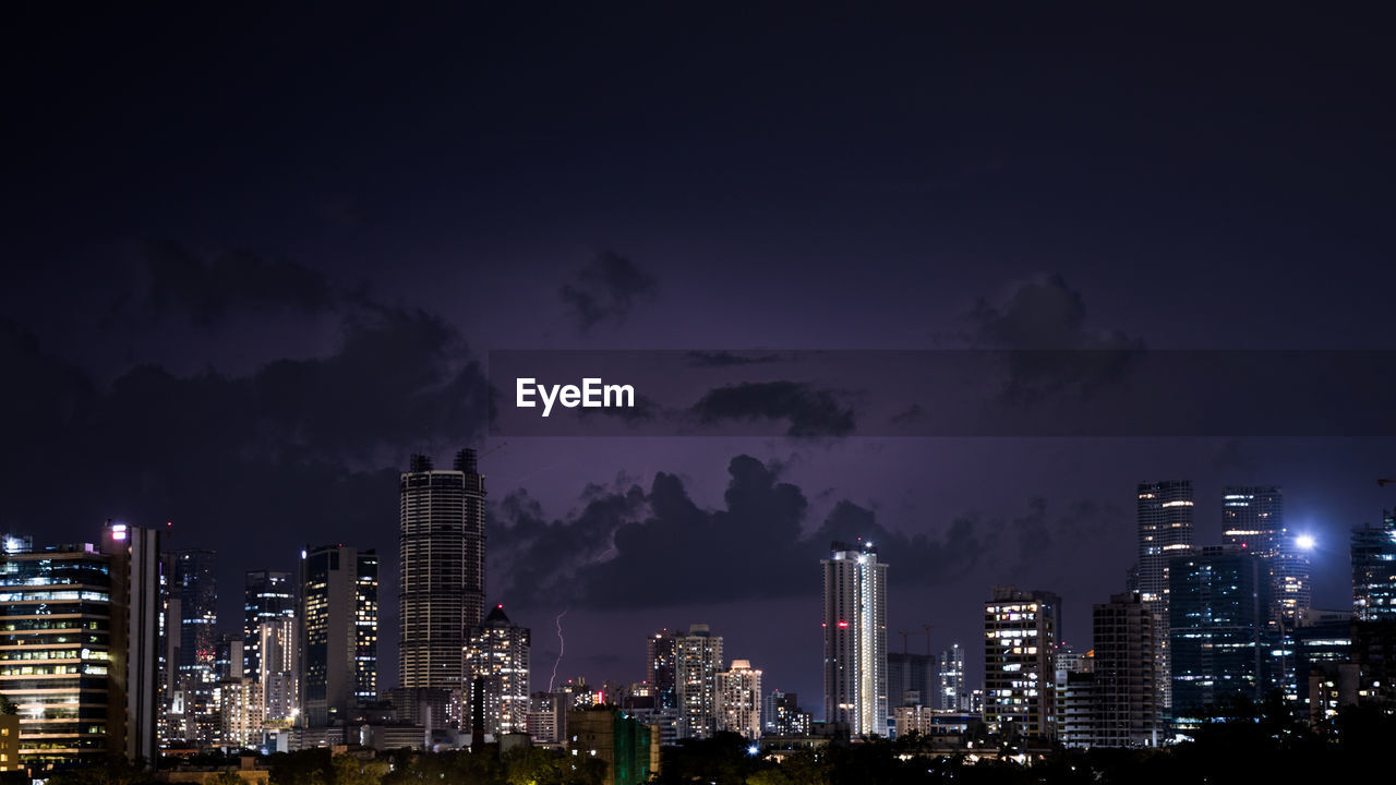 Illuminated buildings and lightning in city against sky at night