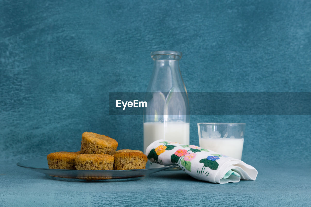 Small cakes in glass plate, with embroidered napkin, milk glass and bottle on blue background