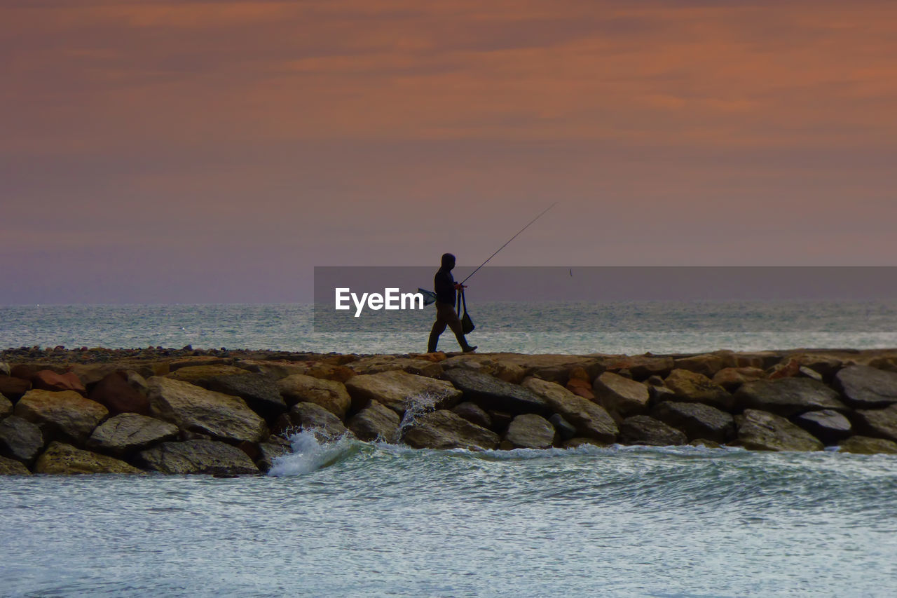 Silhouette man fishing in sea against sky during sunset