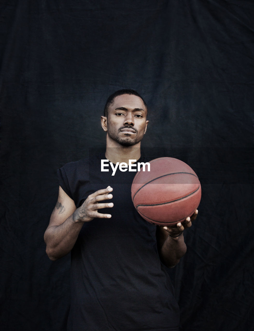 Portrait of man holding basketball while standing against black wall