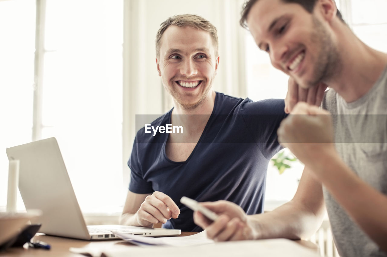 Happy loving homosexual couple with laptop siting at table in home