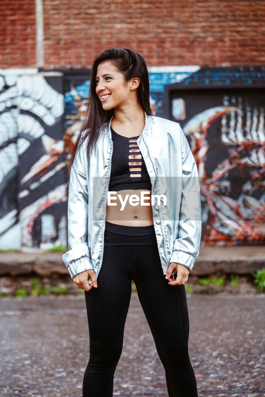 FULL LENGTH OF SMILING WOMAN STANDING AGAINST WALL