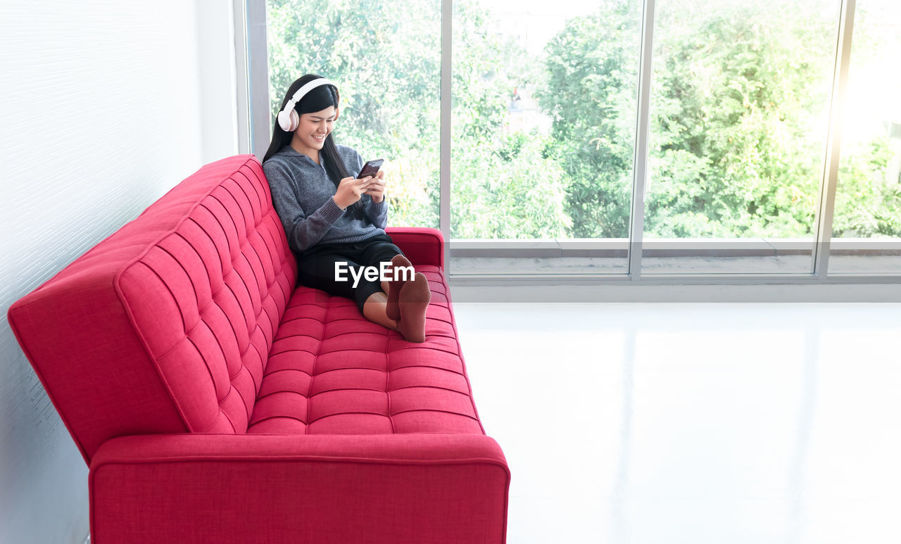 YOUNG WOMAN USING PHONE WHILE SITTING ON SOFA