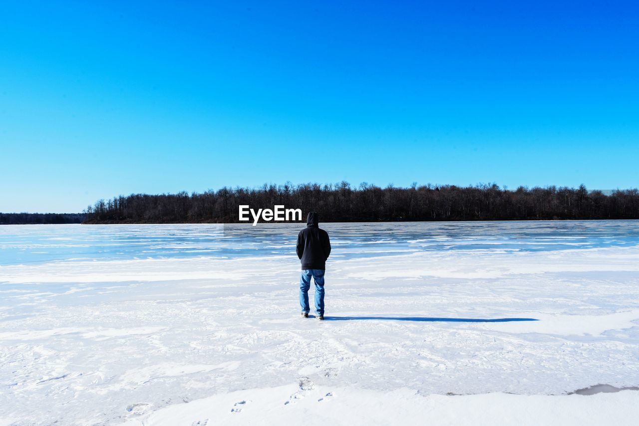 Rear view of man looking at frozen lake against clear blue sky
