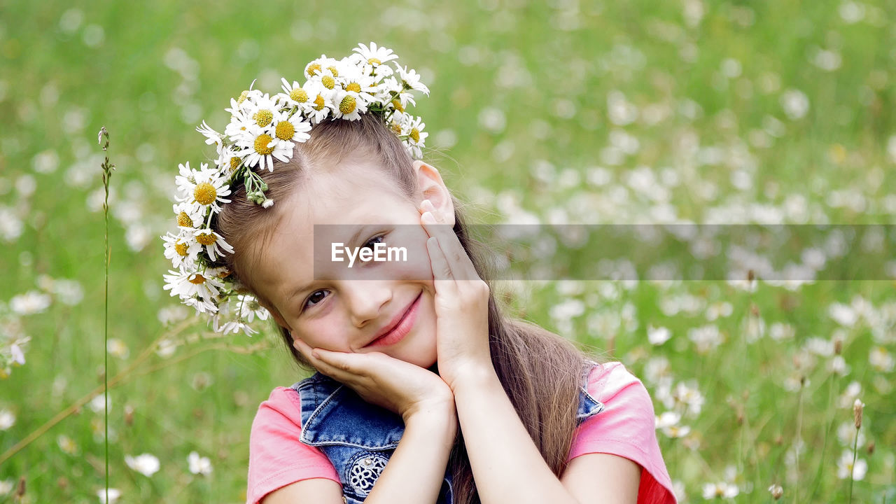 On a chamomile lawn, a sweet girl in a wreath of daisies, smiling, pressing her hands to her cheeks