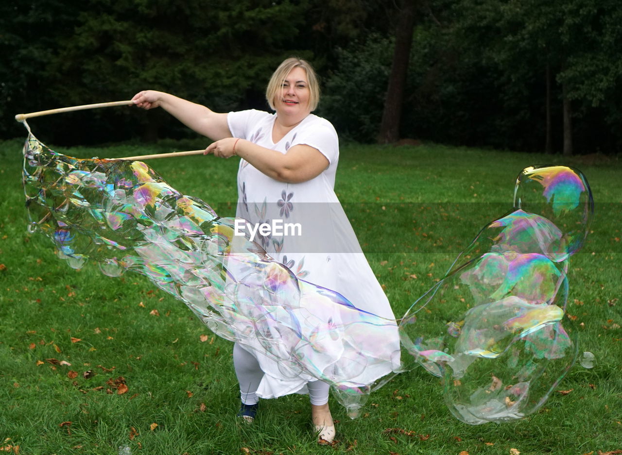 Portrait of smiling woman making bubbles while standing against trees