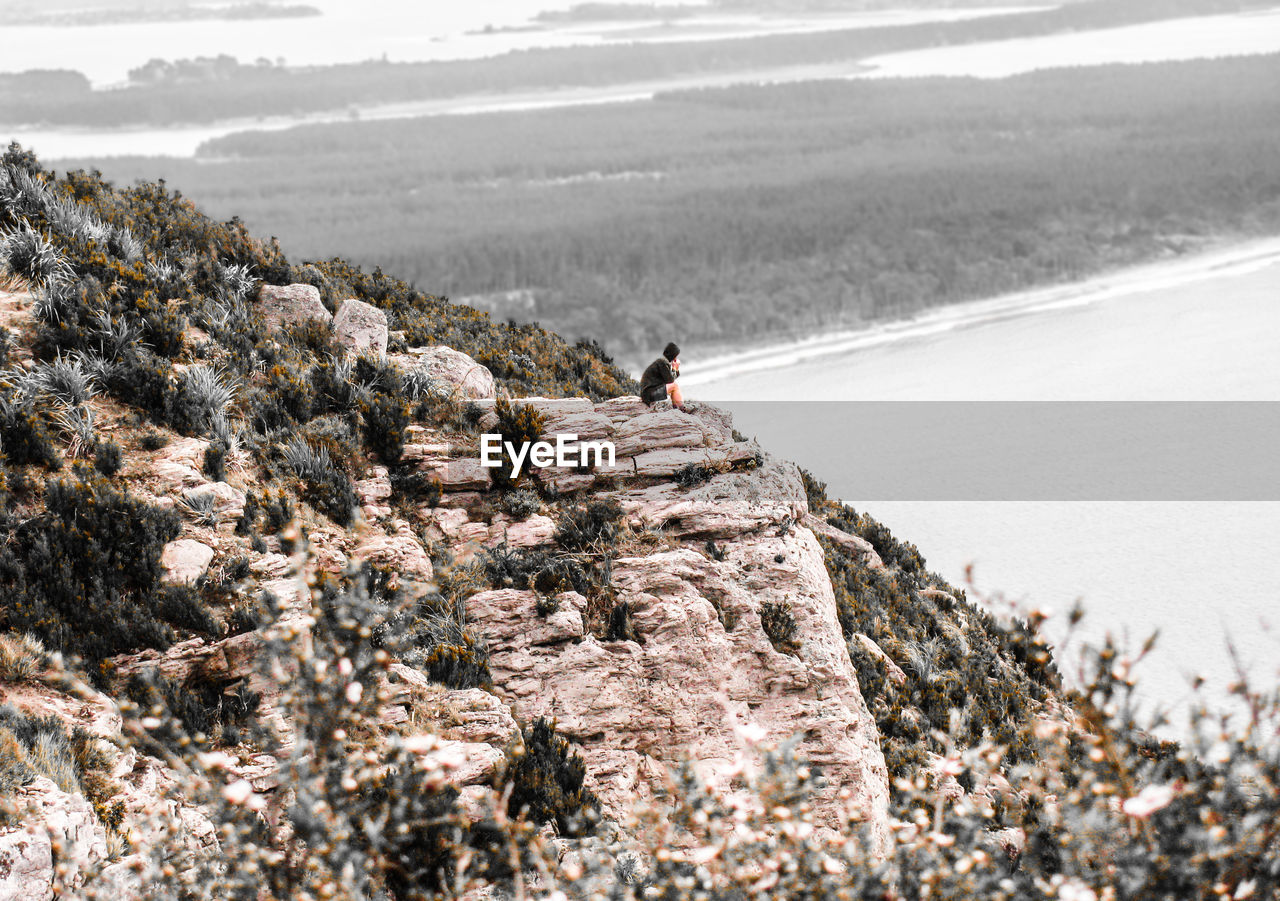 Distant view of man sitting on cliff against river