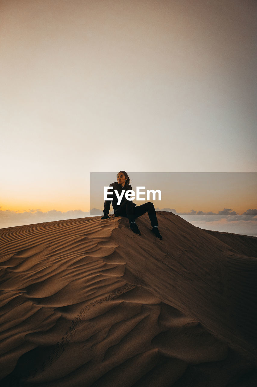 Man sitting on sand in desert against sky during sunrise