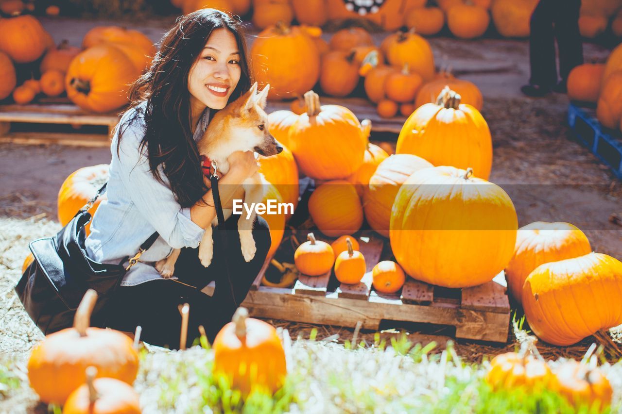 Portrait of smiling young woman with dog crouching amidst pumpkins during halloween