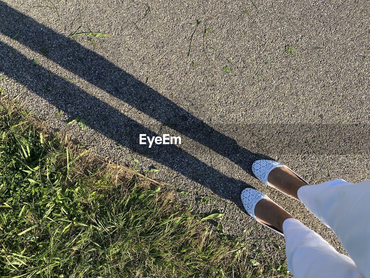 high angle view, human leg, low section, day, grass, one person, nature, sunlight, personal perspective, plant, green, shadow, shoe, lifestyles, outdoors, road, standing, leisure activity, field, men, land, limb, human limb, lawn, adult, flooring