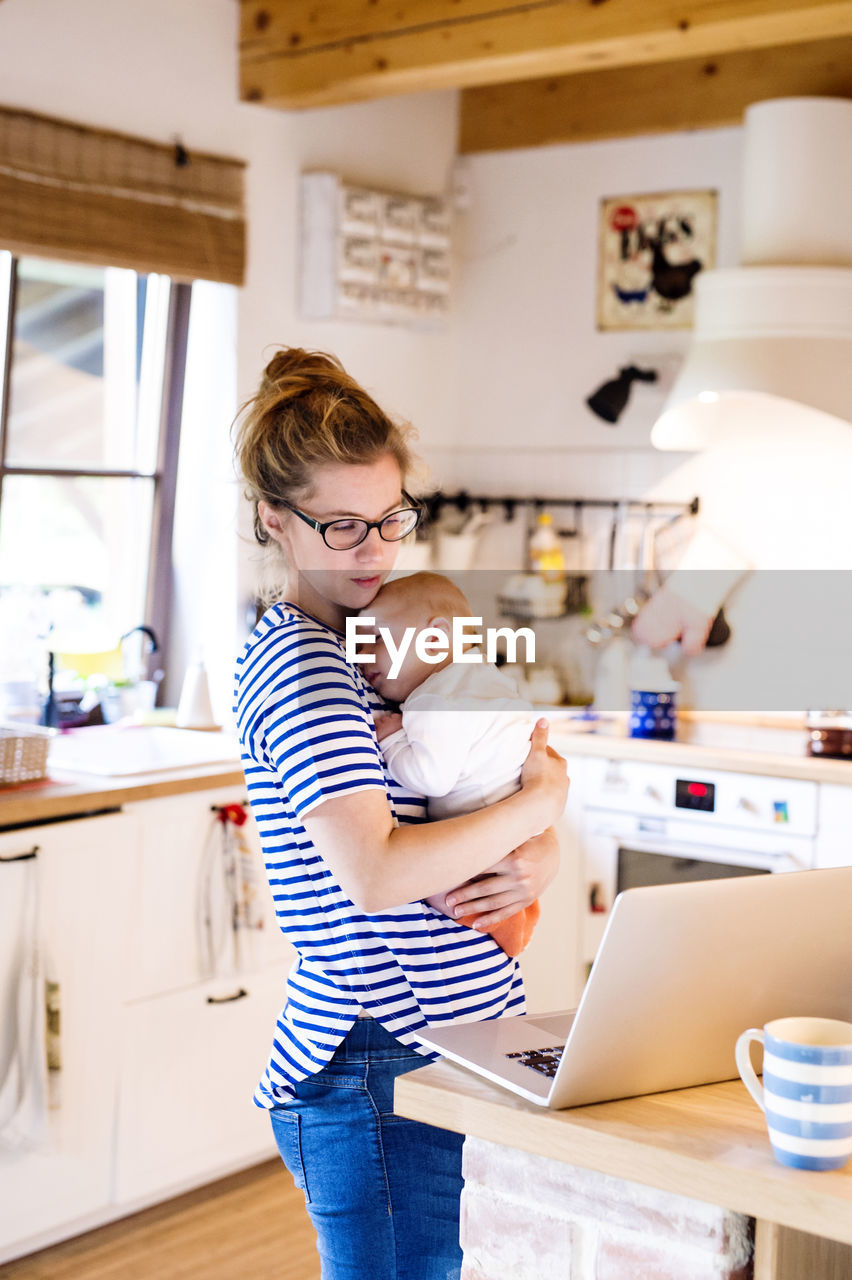 Mother with baby in kitchen looking at laptop