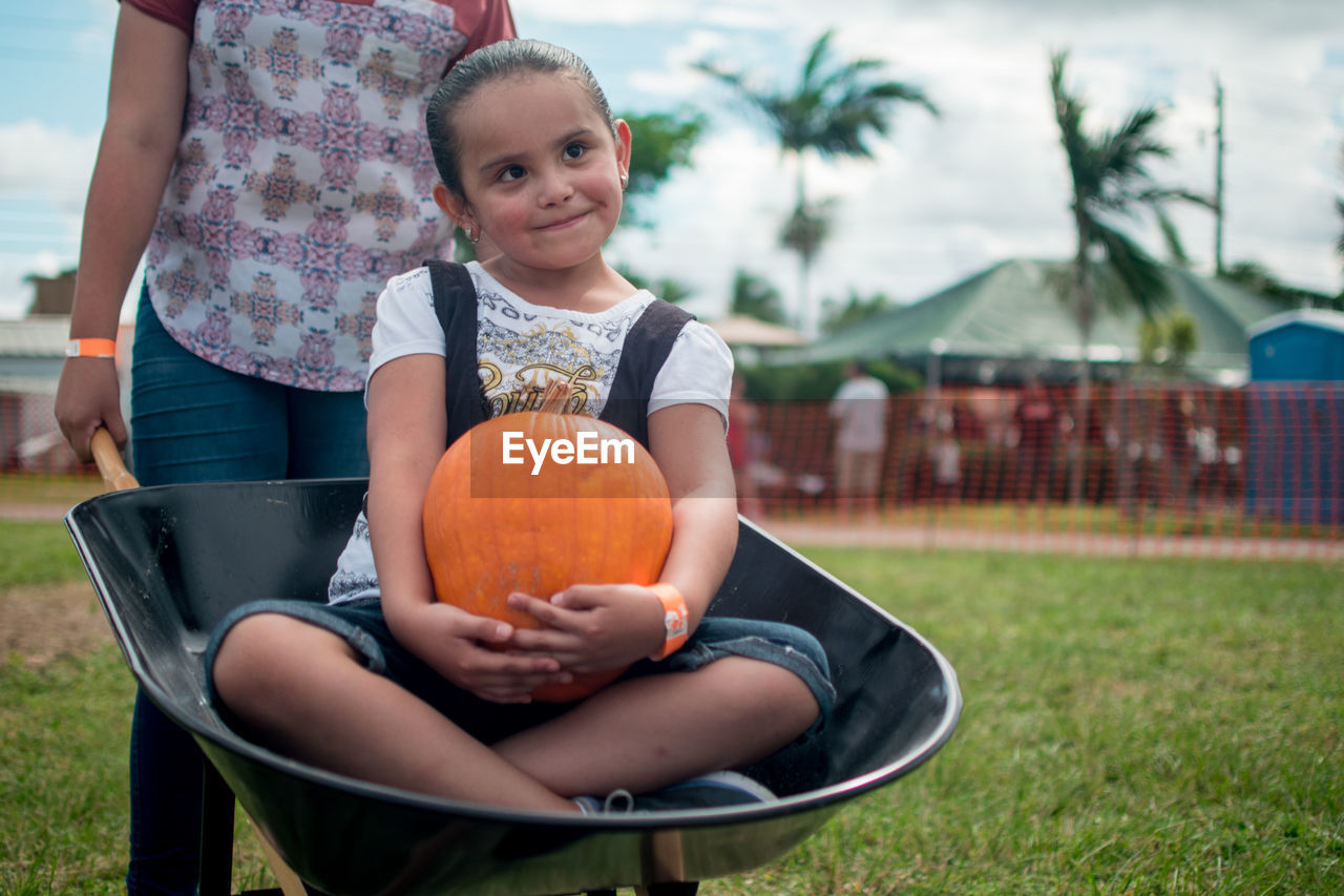 Cute girl with pumpkin sitting in wheelbarrow