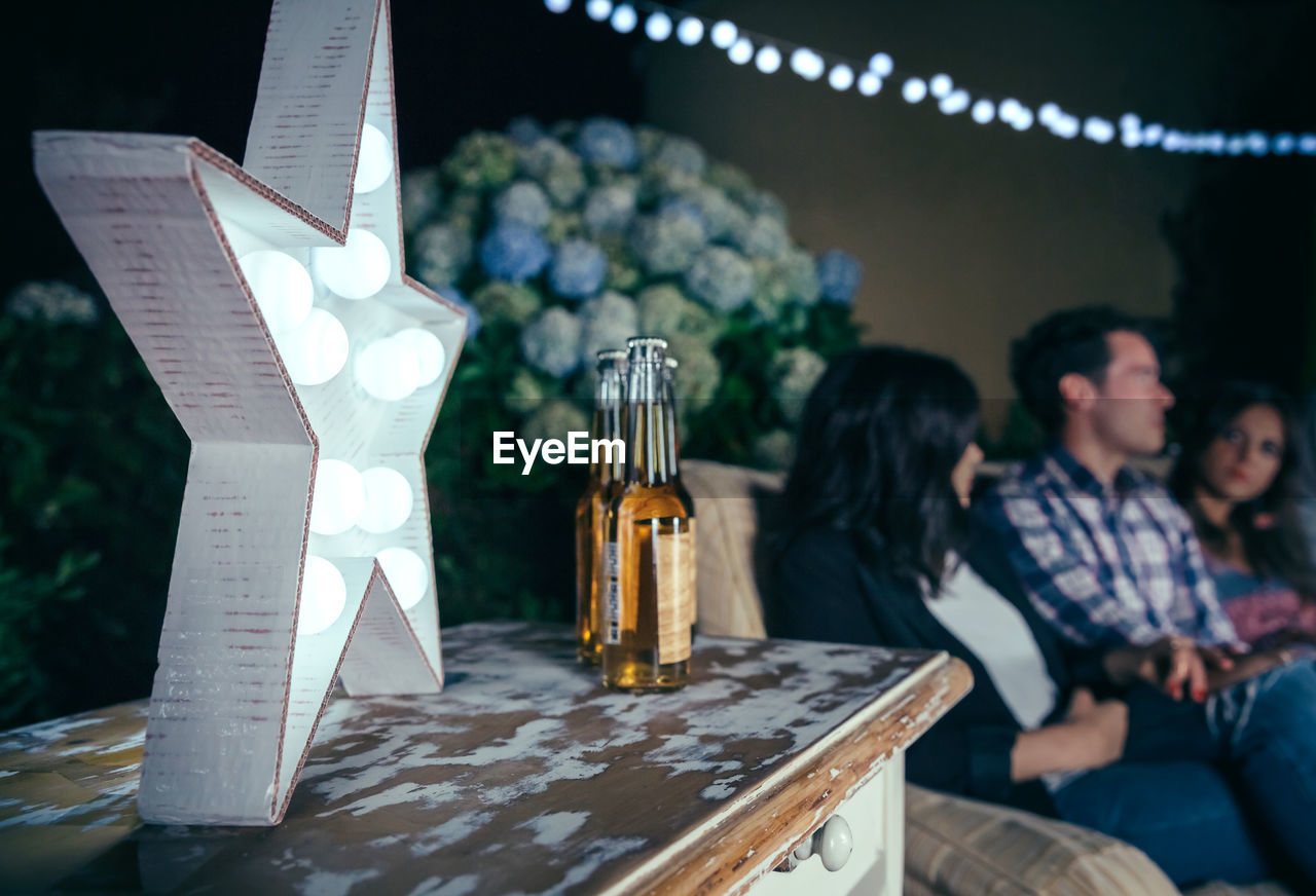Star shape lighting equipment by beer bottles on table with friends in background at night