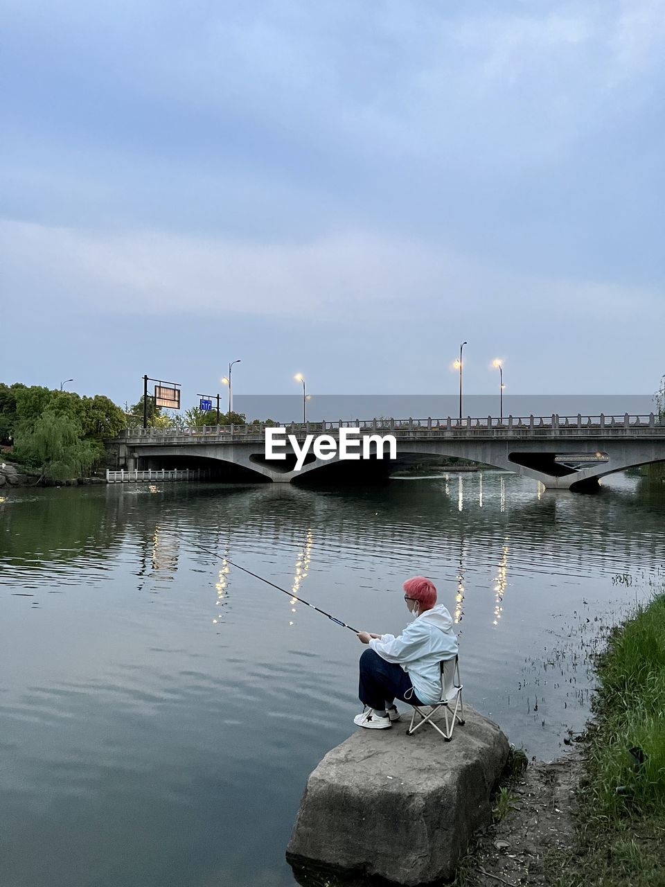 water, river, architecture, sky, nature, built structure, transportation, sitting, men, one person, day, boating, adult, reflection, nautical vessel, shore, full length, reservoir, cloud, outdoors, waterway, bridge, pier, building exterior, occupation, lifestyles, bay, vehicle, rear view, environment, fishing, beauty in nature, tranquility