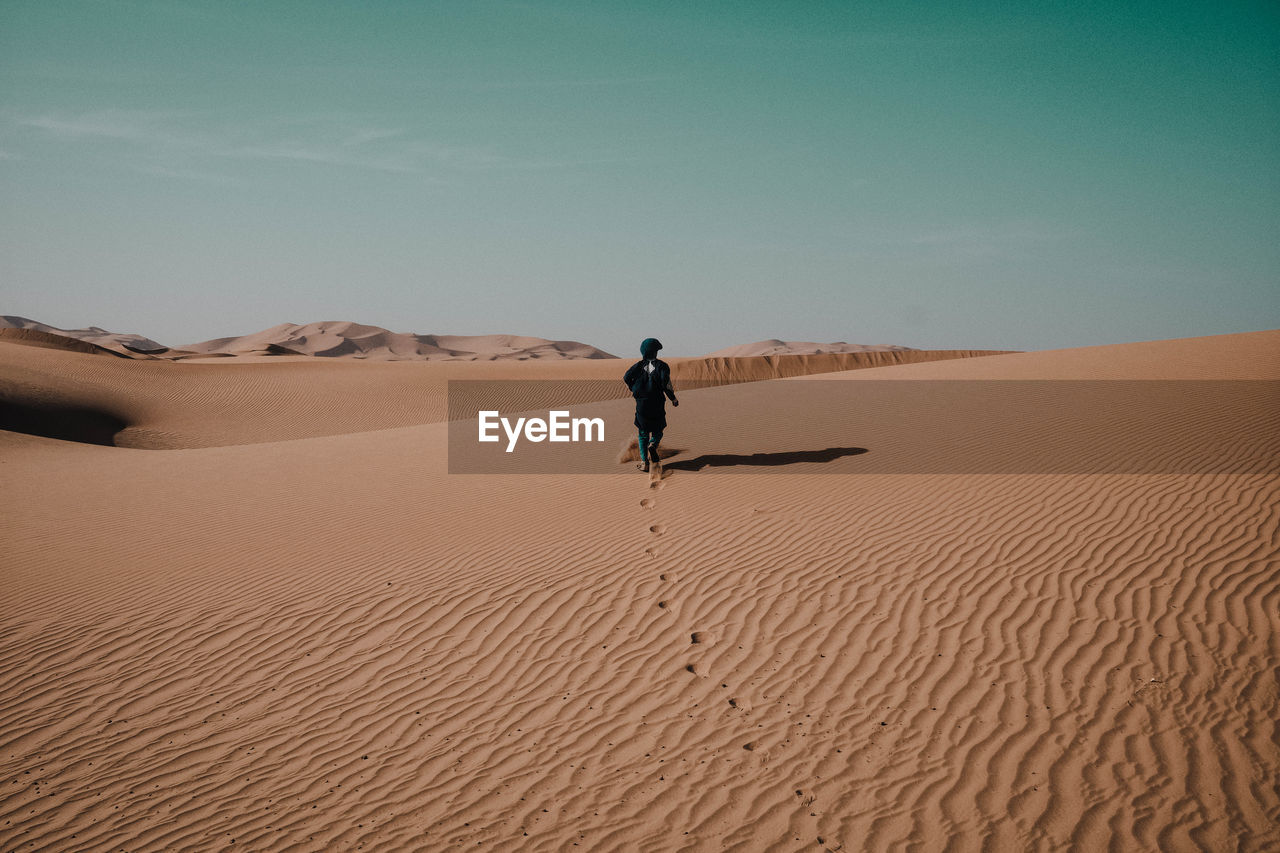 Rear view of man walking on sand at desert against clear sky