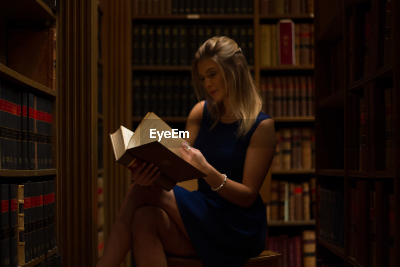 Woman reading book while sitting on stool in library