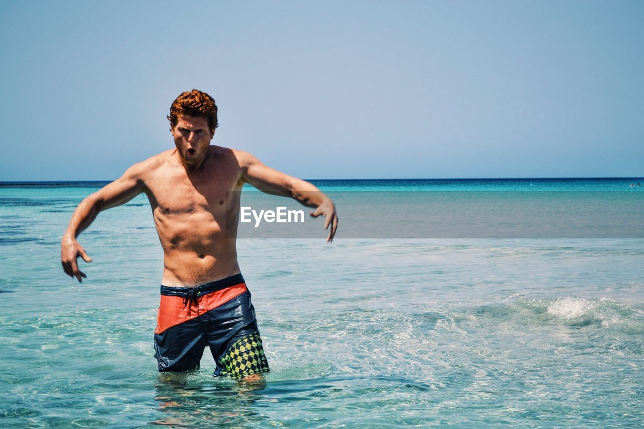 Portrait of shirtless man making face in sea against sky on sunny day