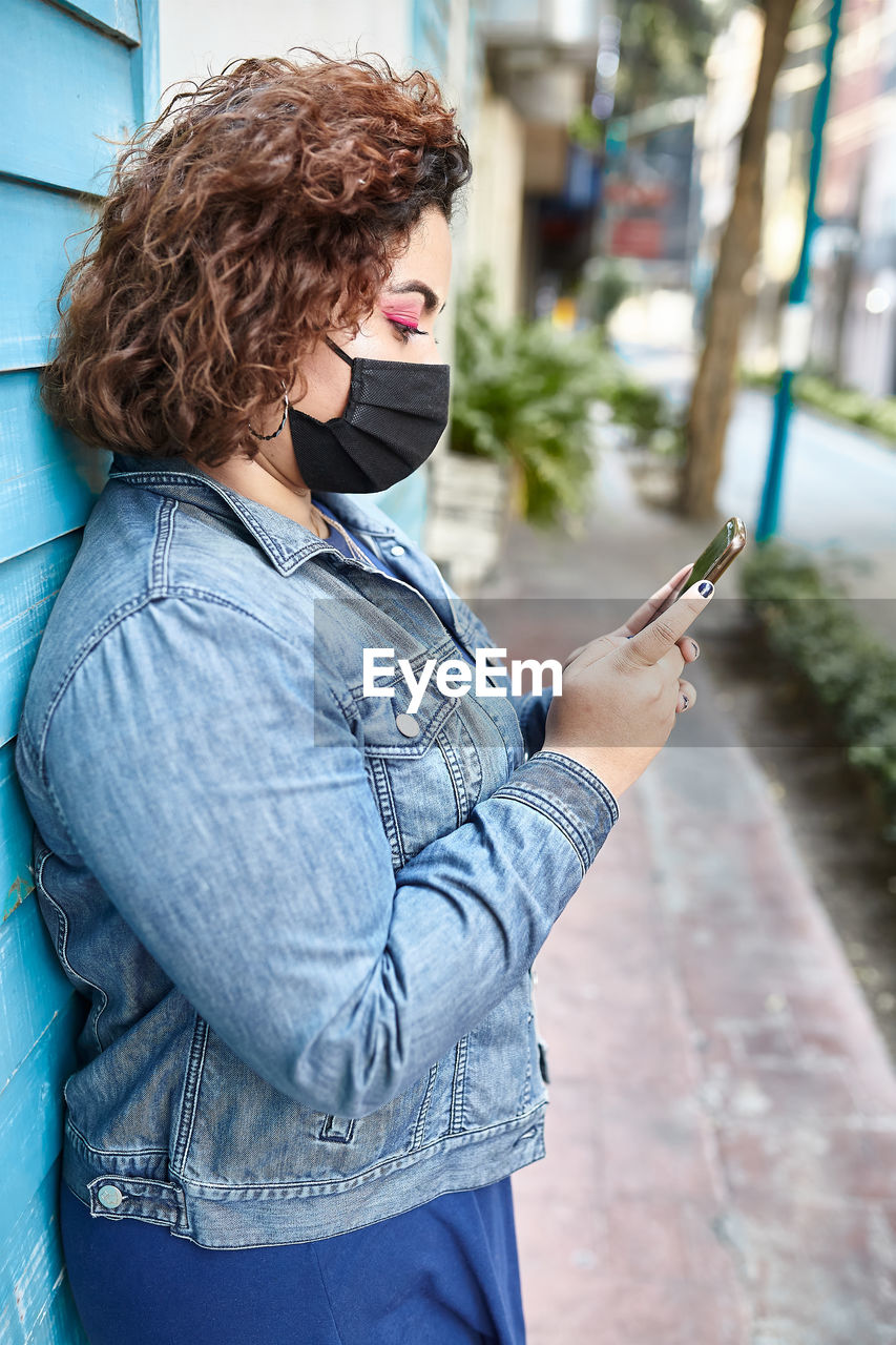 Side view of plus size female wearing protective mask standing in narrow street near residential building and browsing smartphone during covid 19 epidemic in city