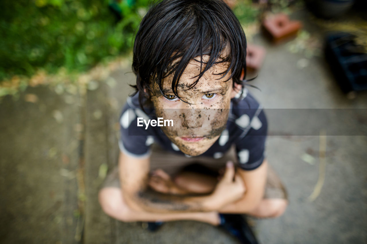 High angle portrait boy with dirty face sitting on footpath at yard during rainy season