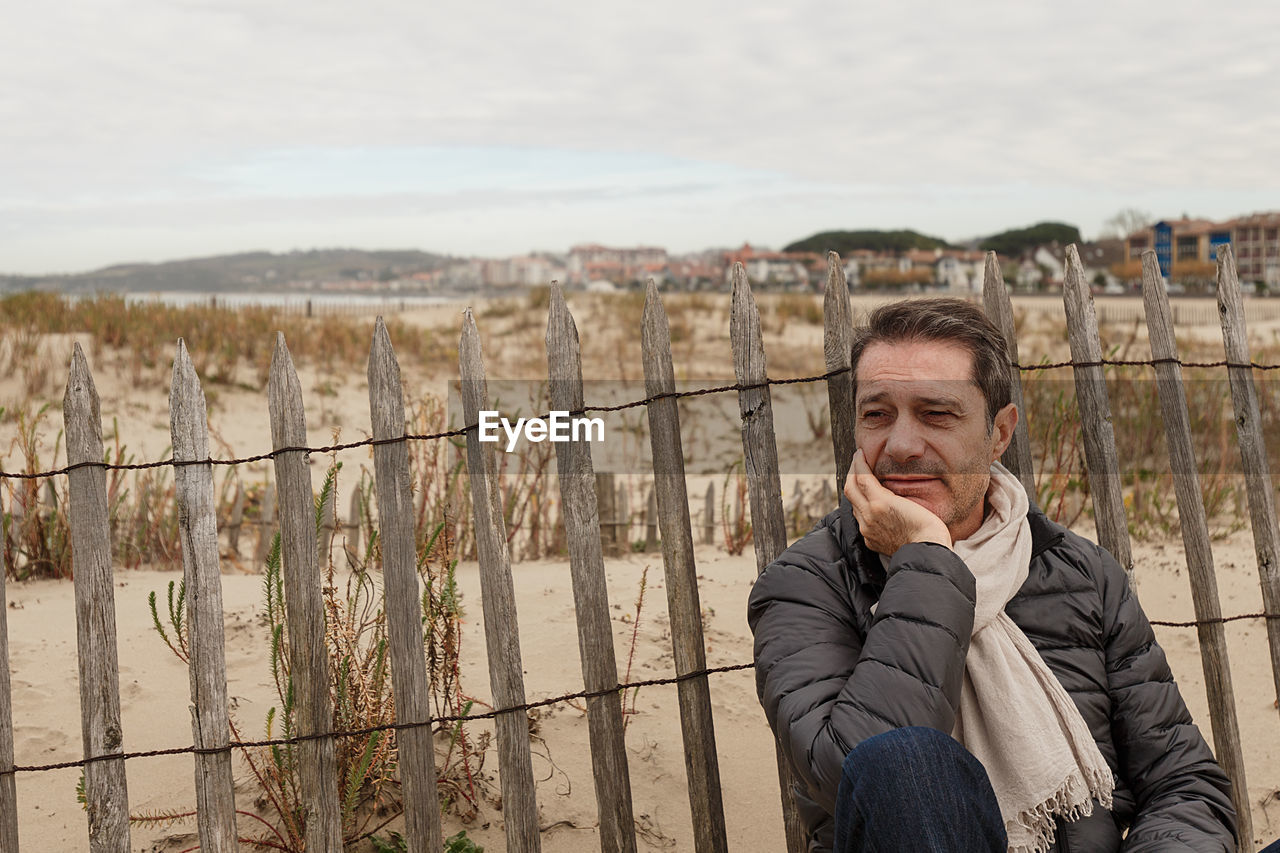 Man looking away while sitting by fence against sky