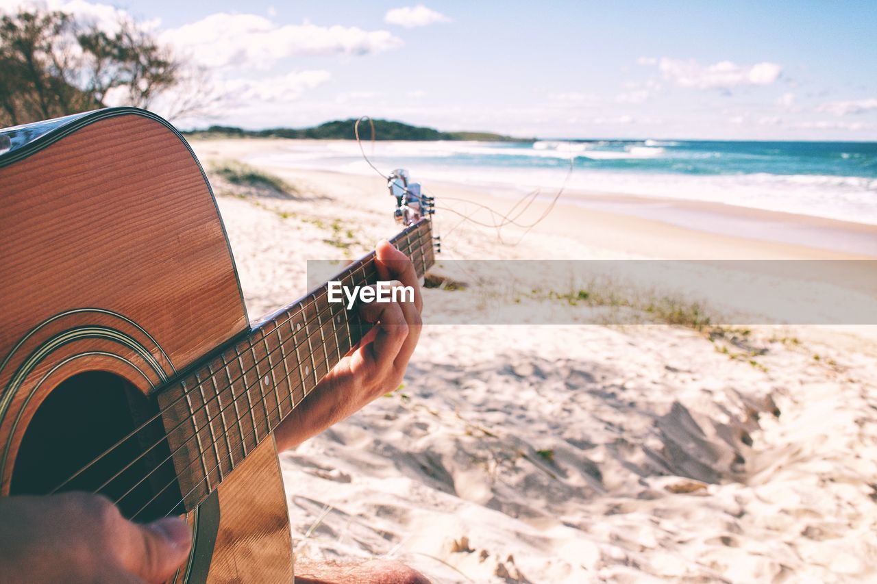 Cropped hand of man playing guitar at beach against sky