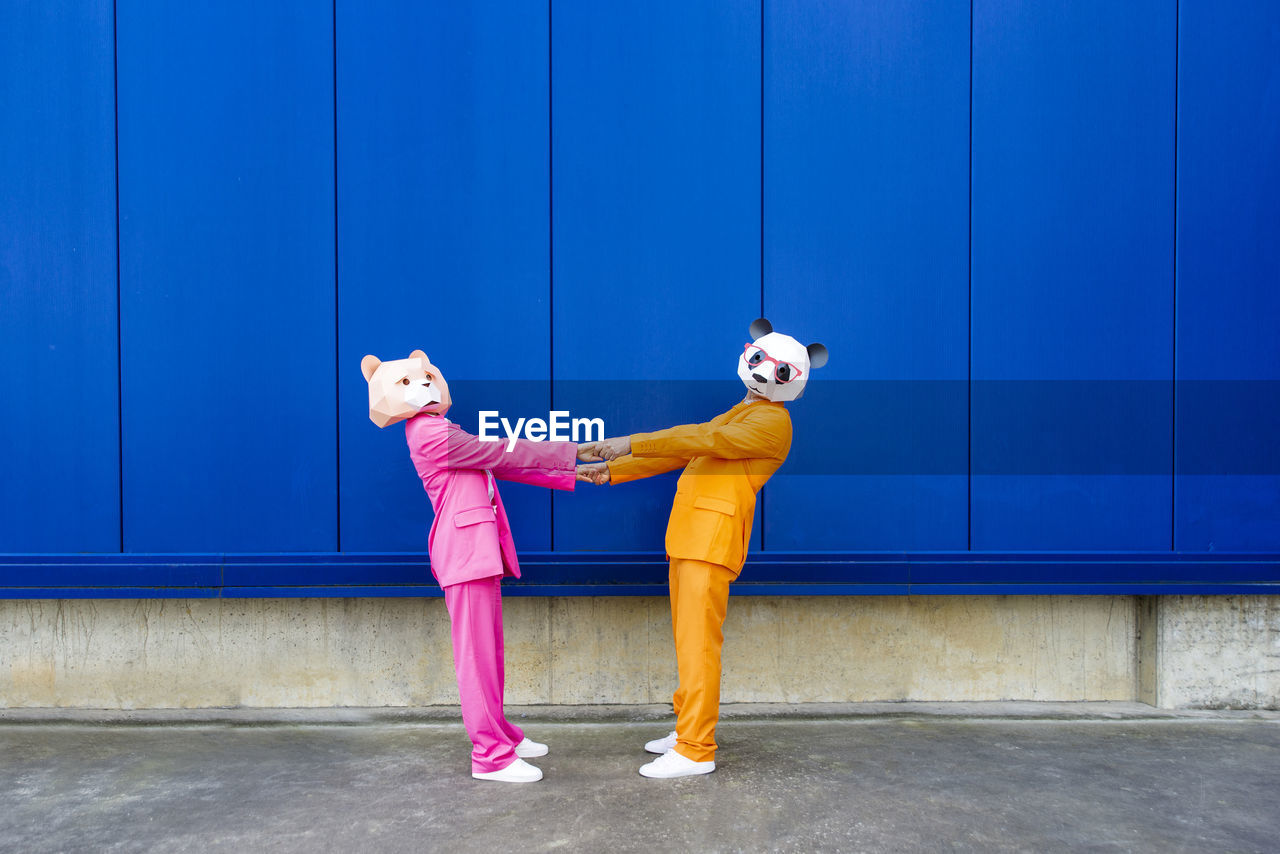 Man and woman wearing vibrant suits and bear masks holding hands against blue wall