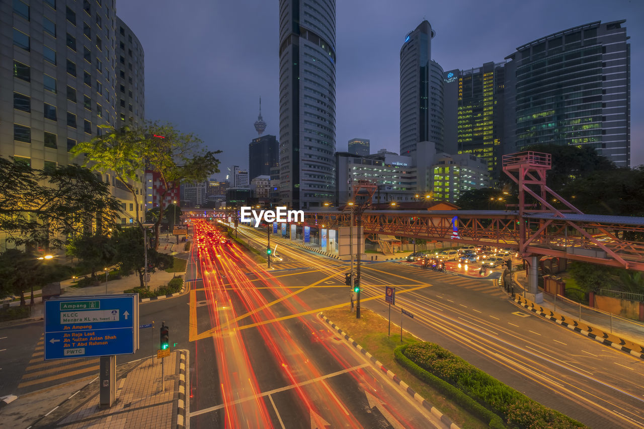 Light trails on city street by buildings against sky