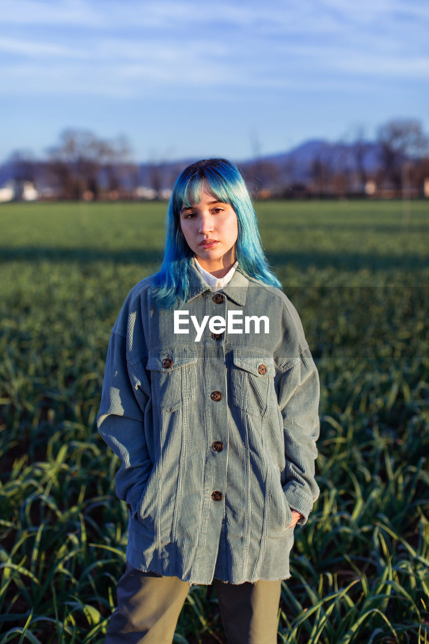 Thoughtful young female with blue hair looking at camera dressed in trendy jacket standing in green field in sunny evening