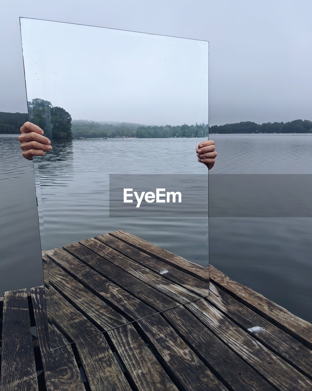 Cropped hand holding mirror on pier at lake against sky