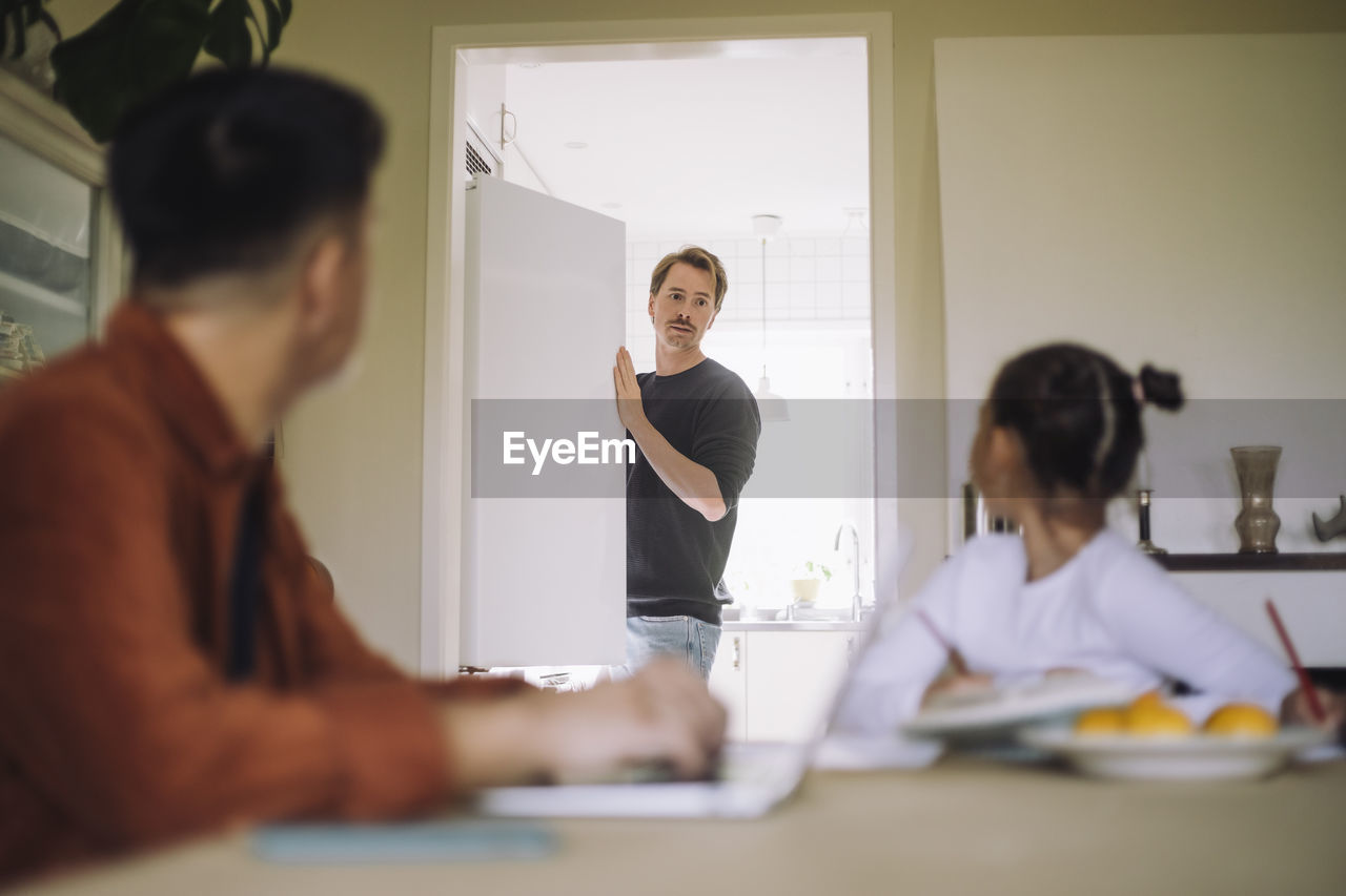 Daughter looking at father standing near refrigerator in kitchen at home