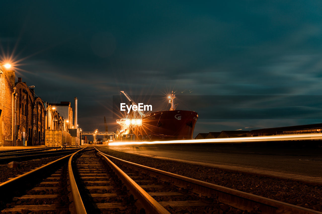 Light trails by railroad tracks at harbor against sky at dusk
