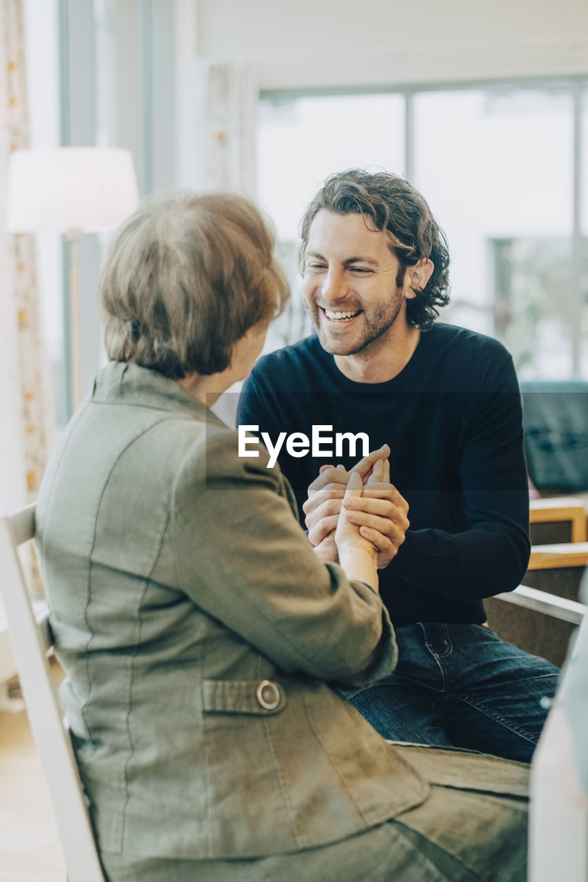 Smiling man holding hands while talking to grandmother at elderly nursing home
