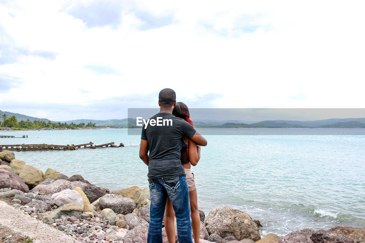 Rear view of man and woman standing by river against cloudy sky