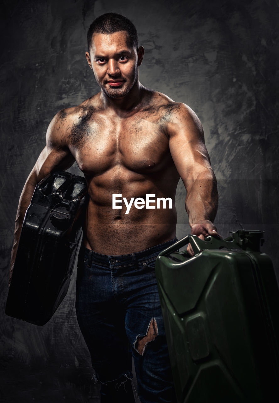 Portrait of shirtless muscular worker holding cans while standing against wall