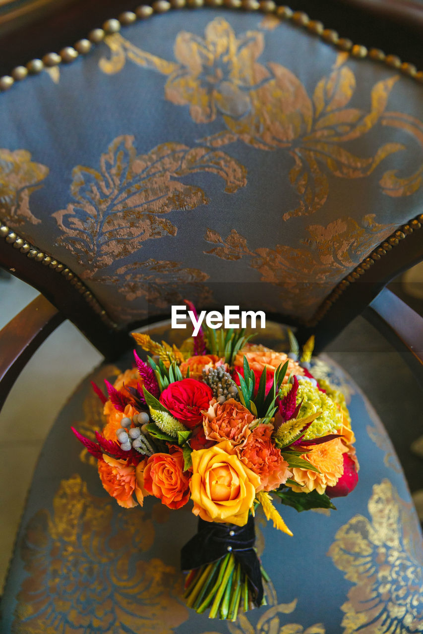 A bouquet of orange roses and alstormeria stands on a dark green chair trimmed with golden leaves