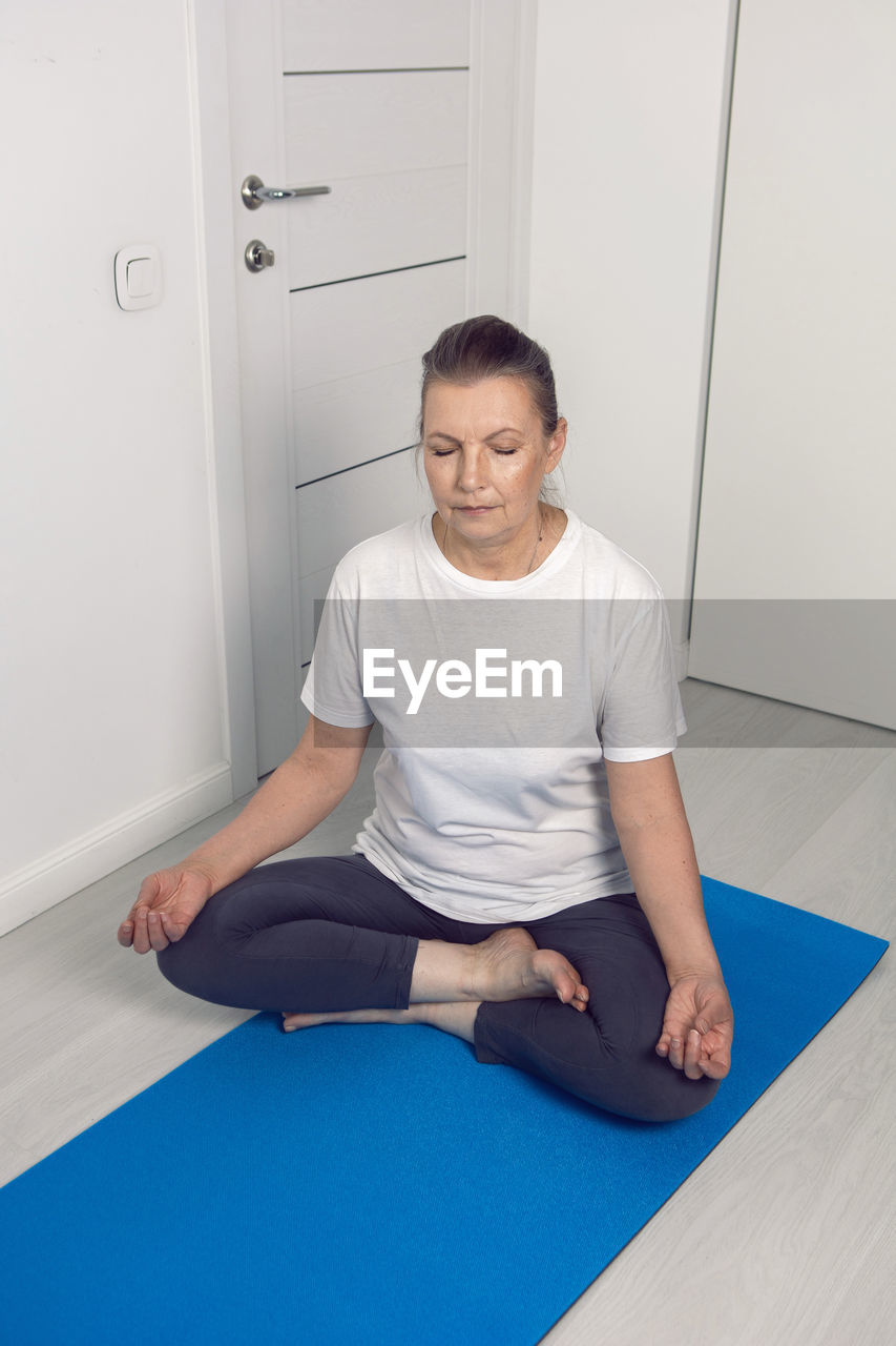 Elderly yoga woman is sitting at home on a yoga mat