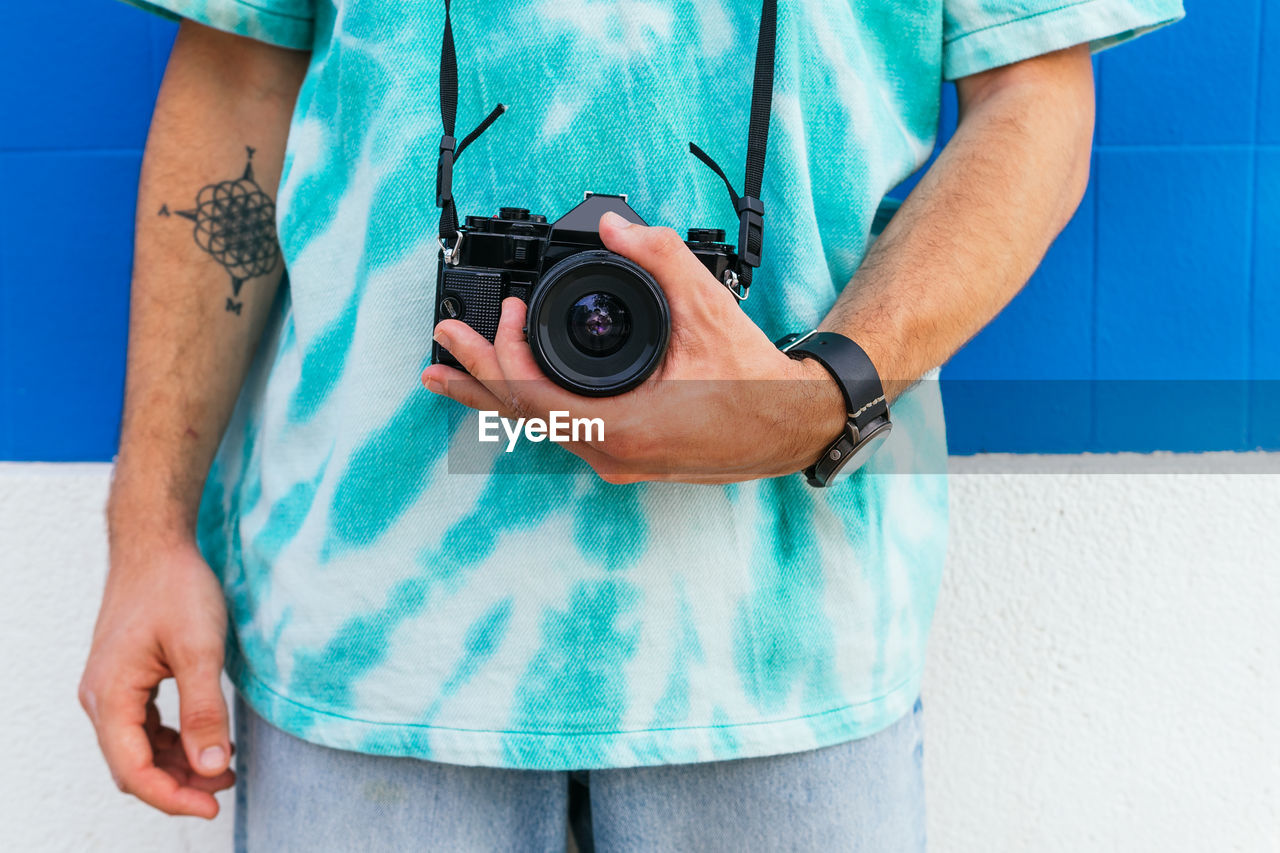 Cropped unrecognizable male photographer in blue t shirt with camera standing on the street outside contemporary blue building wall in city