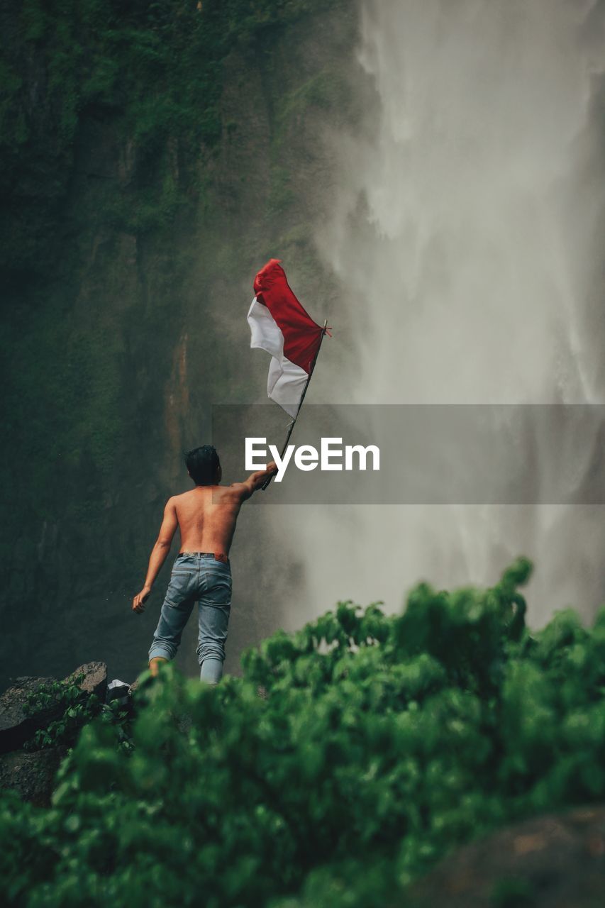 Rear view of man standing by waterfall with indonesian national flag